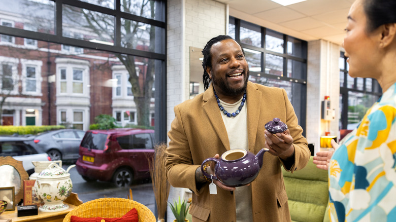 African-American man smiles as he holds a purple teaspot in a thrift or secondhand store while speaking to a lady to his right
