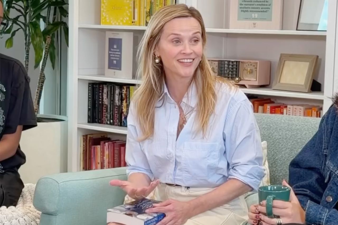Reese Witherspoon sitting in a room filled with books gesturing while talking holding a book in her hand
