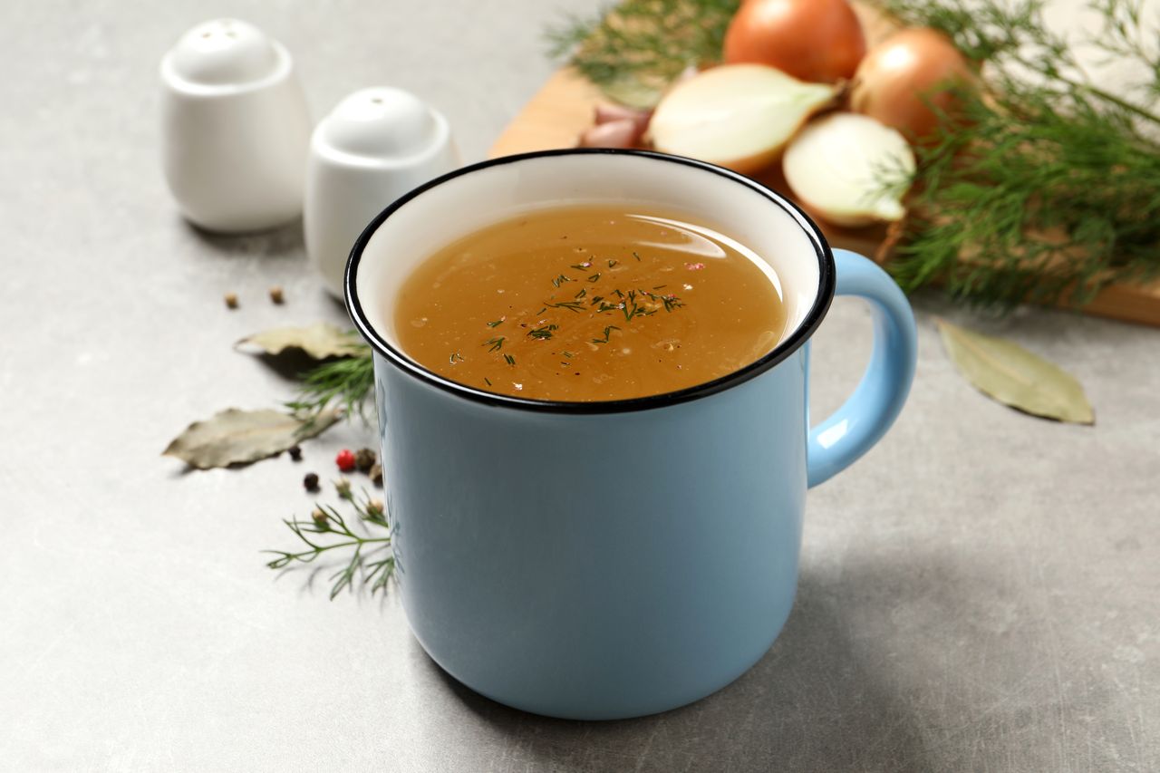 A blue mug filled with broth surrounded by herbs and kitchen ingredients on a surface
