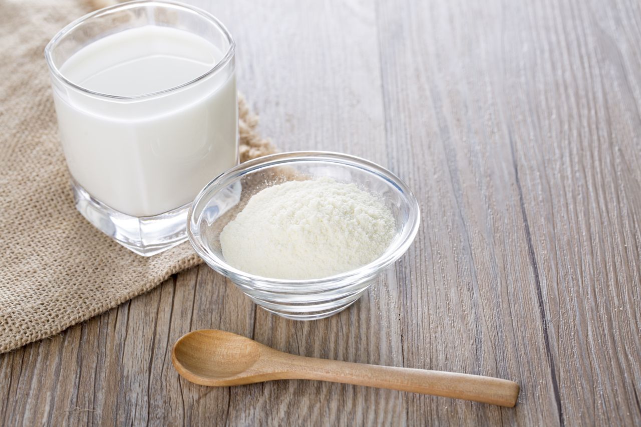 A glass of milk a bowl of powdered milk and a wooden spoon on a wooden surface