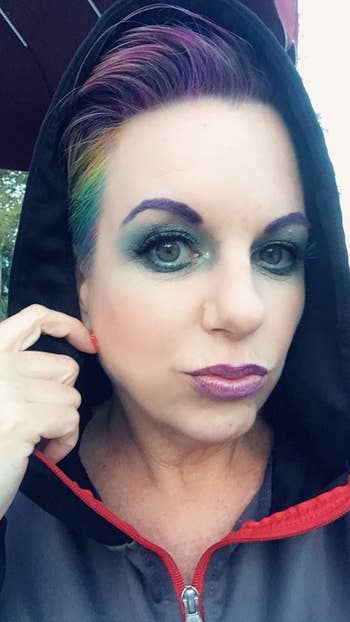 Person with a hooded jacket, colorful short hair, and bold makeup, looking directly at the camera from a close angle