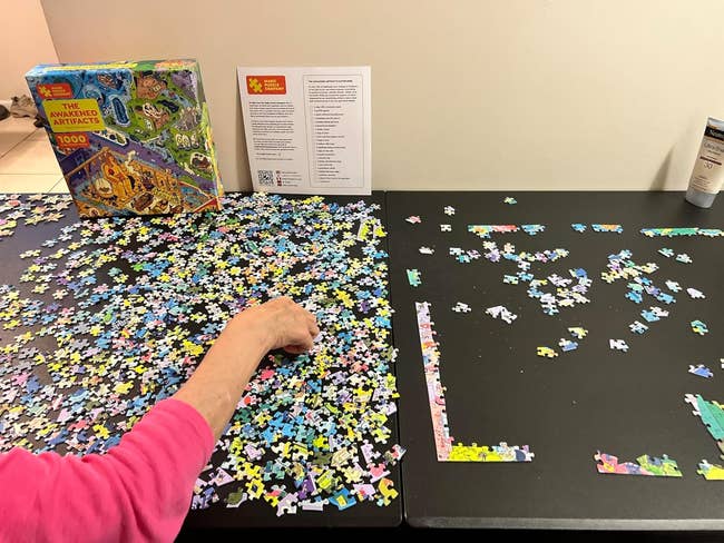 Person assembling a puzzle on a table with scattered pieces, next to a box titled