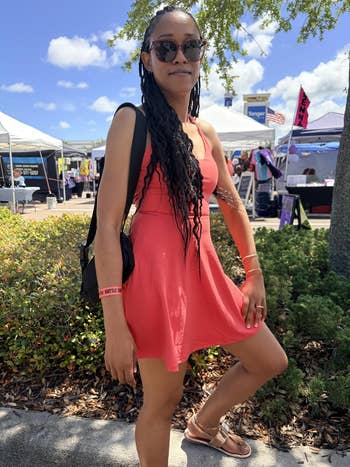 A woman wearing sunglasses and a casual sleeveless dress poses outside at a market event with vendor stalls in the background