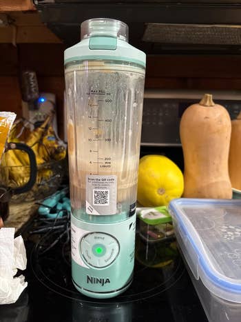 A Ninja blender bottle on a kitchen counter, filled with a smoothie. Surrounding items include fruit, a squash, and a storage container