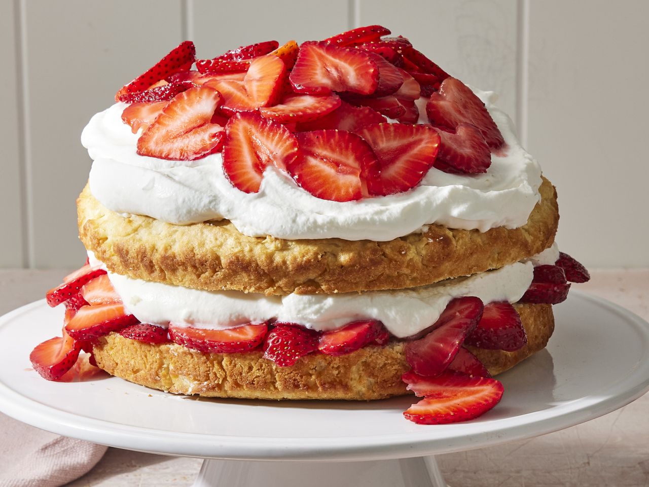 closeup of a large Strawberry Shortcake split and filled with whipped cream and sliced strawberries, topped with more cream and strawberries, on a white cake stand