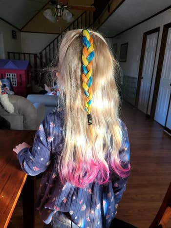 Child with a blue and yellow braid, wearing a floral top, stands with back to camera, showcasing pink-dyed hair tips in a home setting