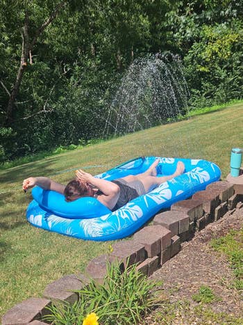 Person relaxing outdoors on a blue inflatable lounger with a water feature spraying overhead, enjoying a sunny day in a grassy backyard