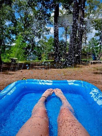 Person's legs in an inflatable pool with water sprays overhead, surrounded by trees and a basketball hoop in the background