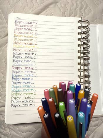 Hand holding colorful marker pens over a notebook, each pen color matches the words “Paper mate” written on the page