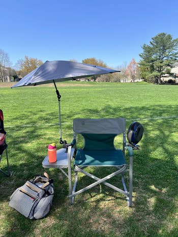 Outdoor setup with a folding chair, umbrella, portable fan, red drink bottle, and a striped bag on grass in a park setting
