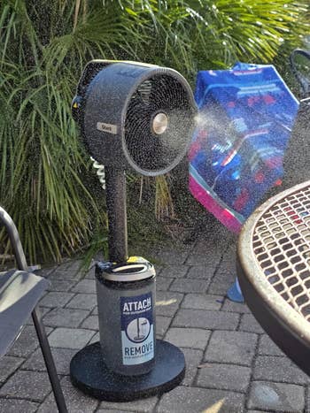 A misting fan sprays water outdoors, next to a blue pool toy. A label on the fan reads