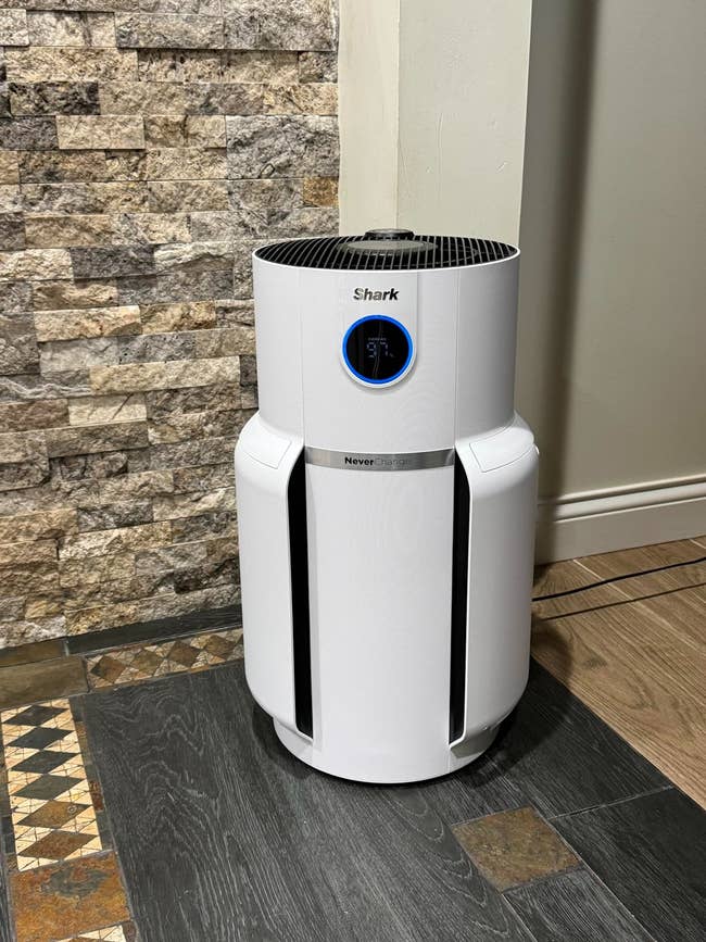Shark air purifier with digital display, placed on a hardwood floor next to a stone wall