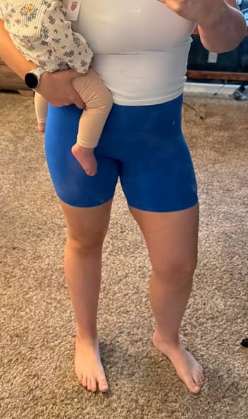 Person in a tank top and blue shorts holds a baby, shown in a casual home setting, suitable for relaxation or lounging