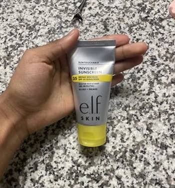 Person holding e.l.f. Skin Suntouchable Invisible Sunscreen SPF 35 tube on a speckled countertop