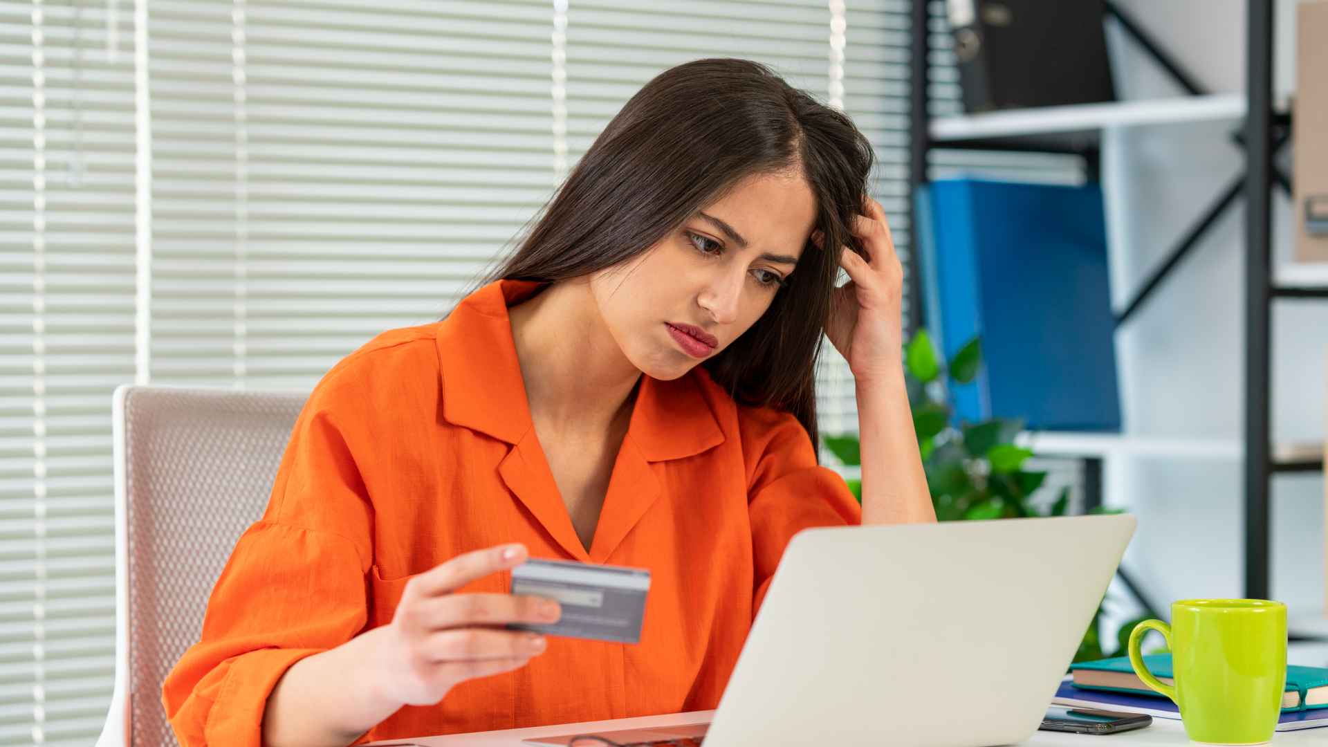 Discover a stressed-looking woman in orange top looking at laptop and holding credit card in right hand