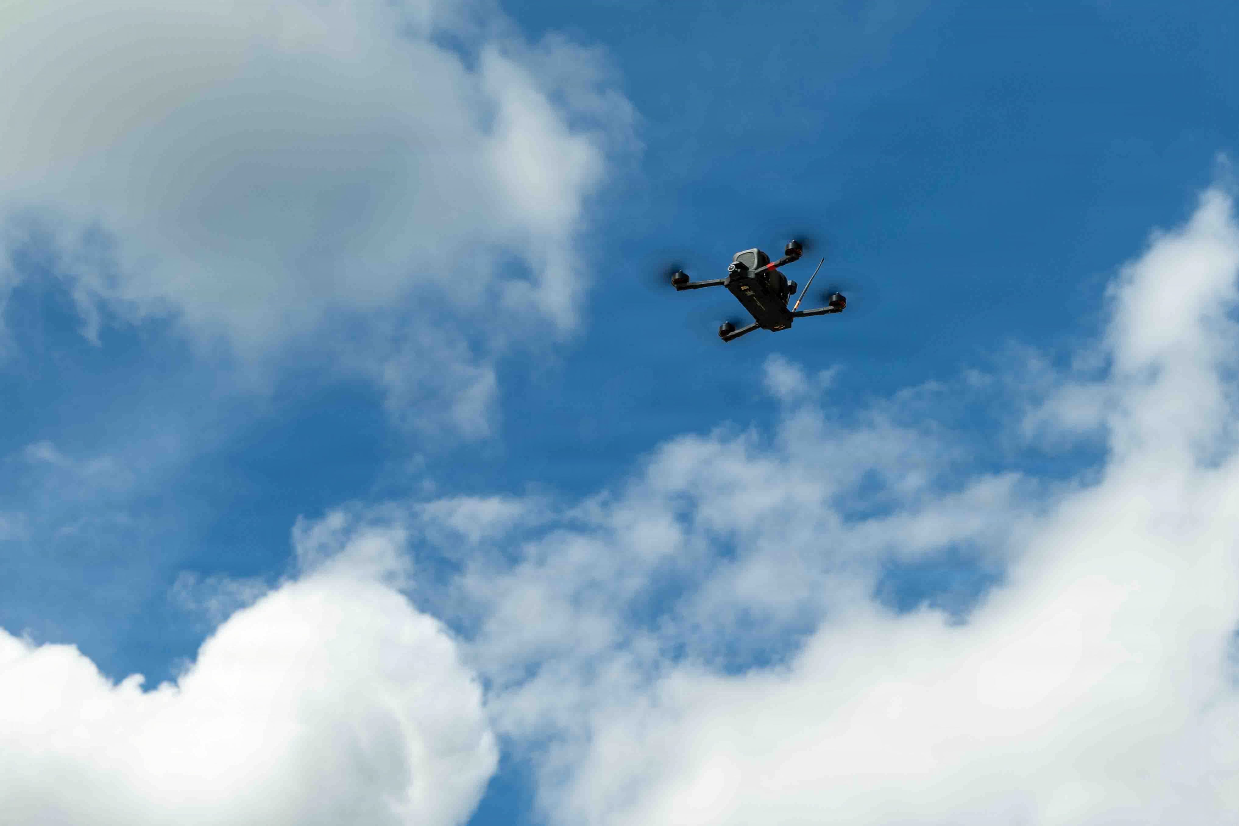 A small black drone flies in a cloudy sky.