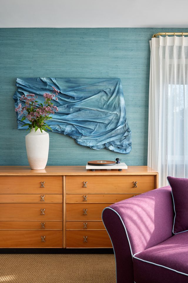A wooden sideboard with a vase of flowers and a turntable wall art of draped fabric on the wall a purple armchair in the foreground