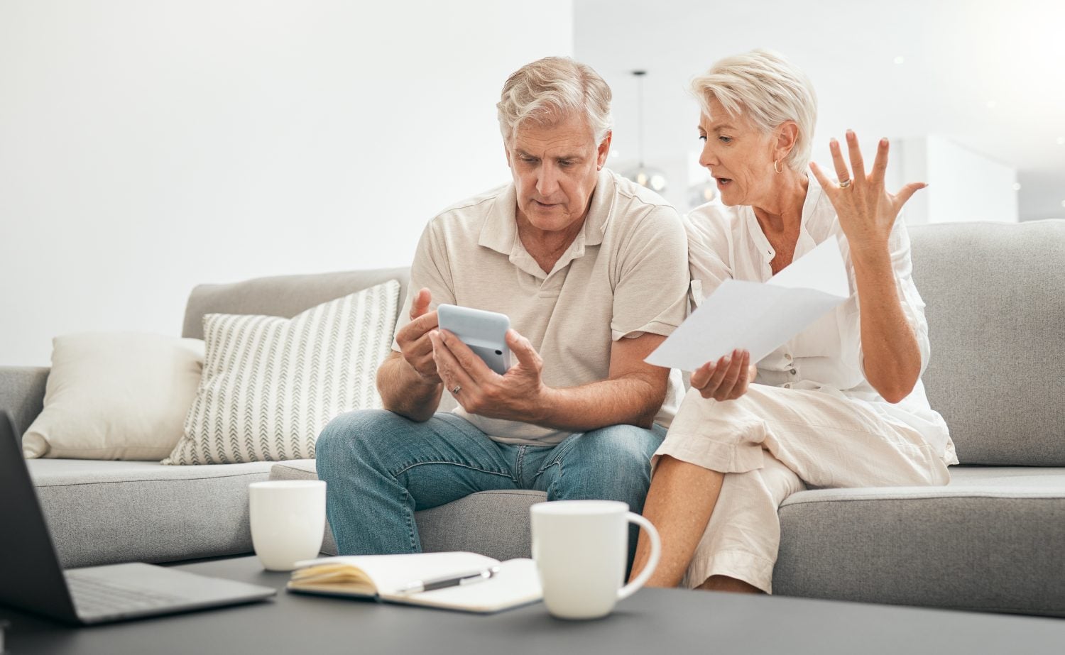 A senior couple sits on a grey sofa in a brightly lit living room, reviewing financial documents. The man, with grey hair, wears a light polo shirt and jeans, intently looking at a white calculator in his hands. The woman, also with grey hair, wears a white blouse and light trousers, holding several white papers and gesturing with her right hand, her expression one of concern or frustration. On a dark coffee table in front of them, there is an open laptop, a notebook with a pen, and two white mugs.