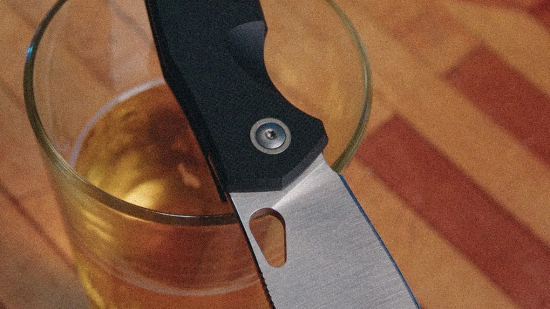 Close-up of a folding knife with a black textured handle resting on the rim of a glass filled with amber liquid.