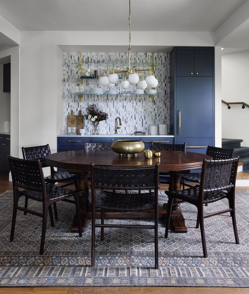 A dining room featuring a round wooden table a set of woven chairs a decorative rug a backsplash blue cabinetry and pendant lighting