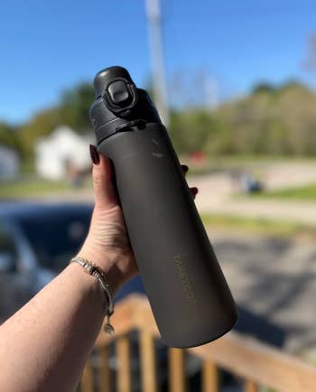 Person holding a matte black TAKETODAY water bottle with a flip-top lid outdoors, showcasing its sleek design