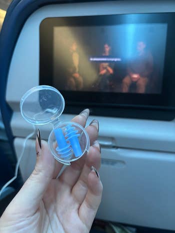 Hand holding earplugs case in front of an airplane seat screen playing a movie