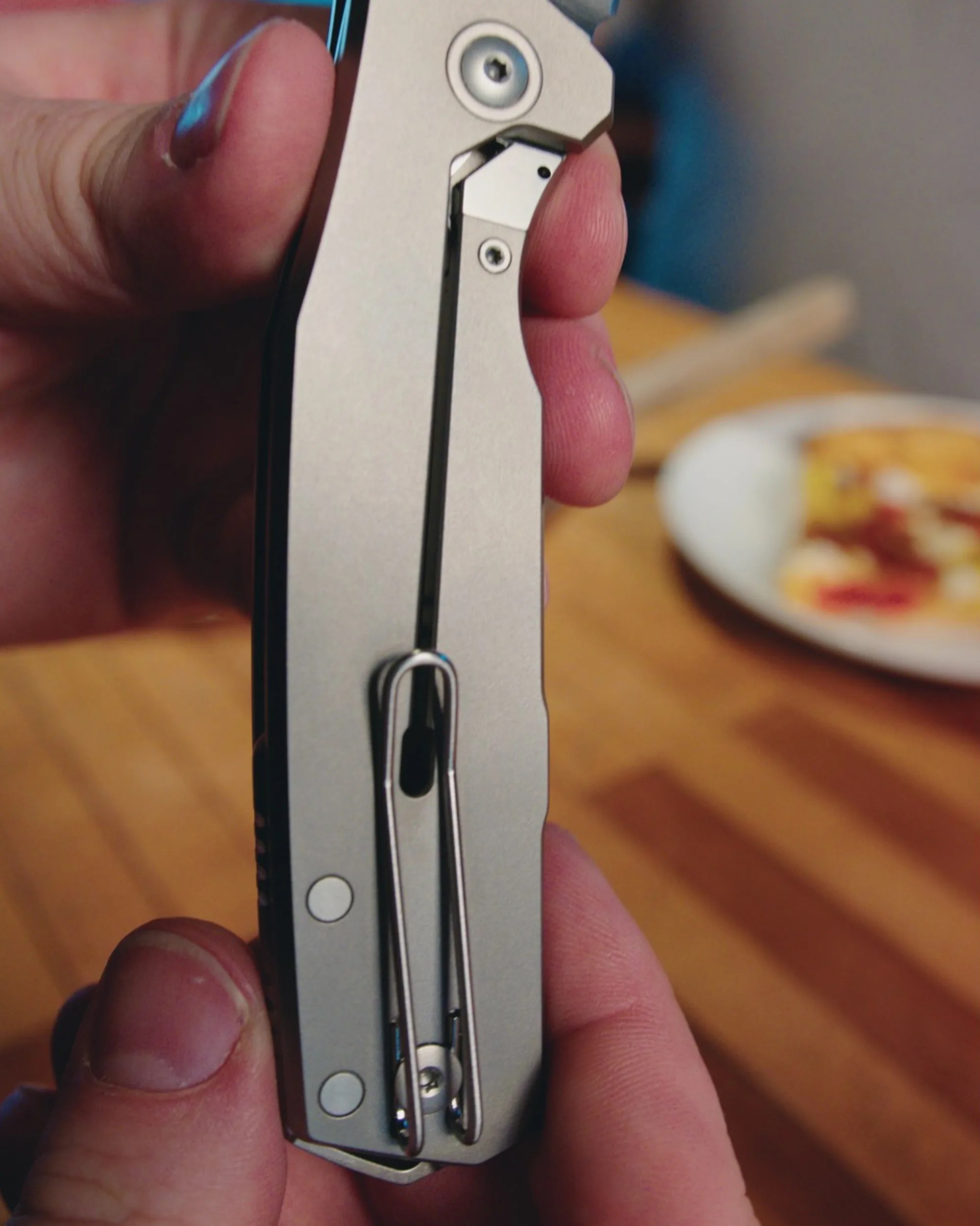 Hand holding a closed silver folding knife with a metal clip on a wooden surface.