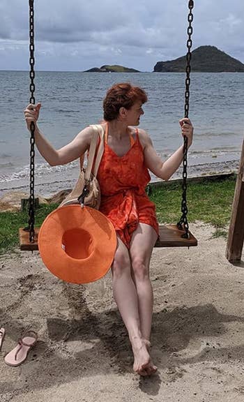 reviewer in a summer dress holding a beach hat and a bag, seated on a swing by the beach with islands in the background