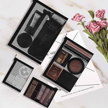 Four transparent zippered pouches filled with makeup products displayed alongside a bouquet and a card on a marble surface