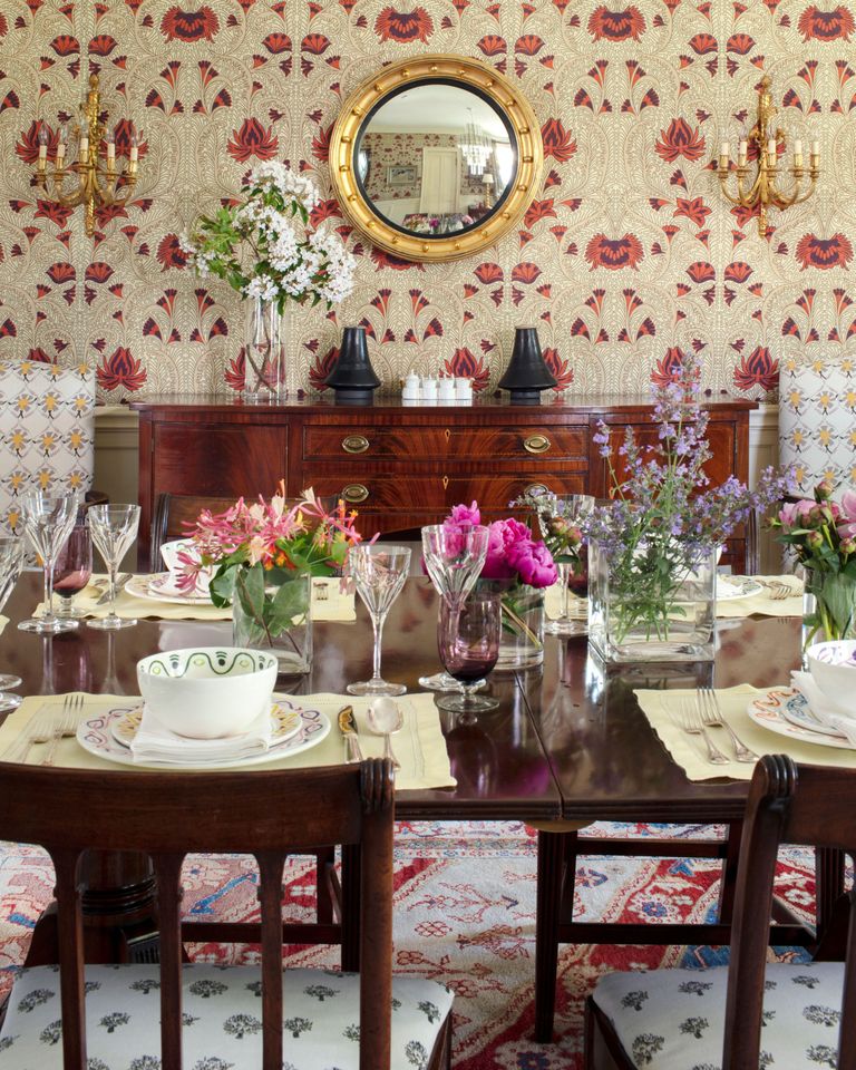 A dining table with floral decorations surrounded by chairs and a sideboard with a mirror and ornaments in the background