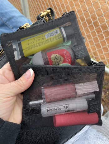Hand holding two mesh pouches filled with assorted lip balms and small beauty products near a chain-link fence