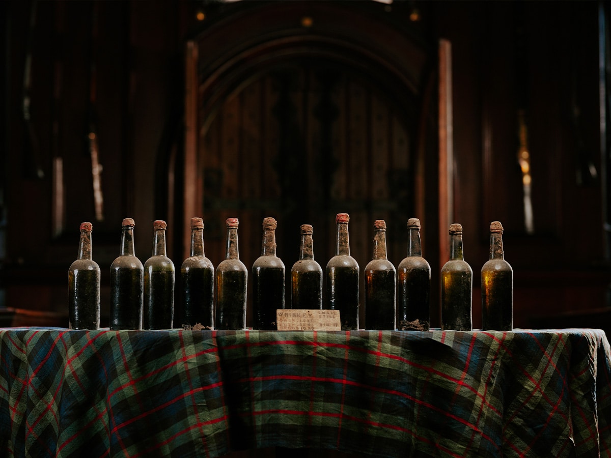 Old whisky bottles lined up on a tartan-covered table at Blair Castle, Scotland, with a wooden door in the background.