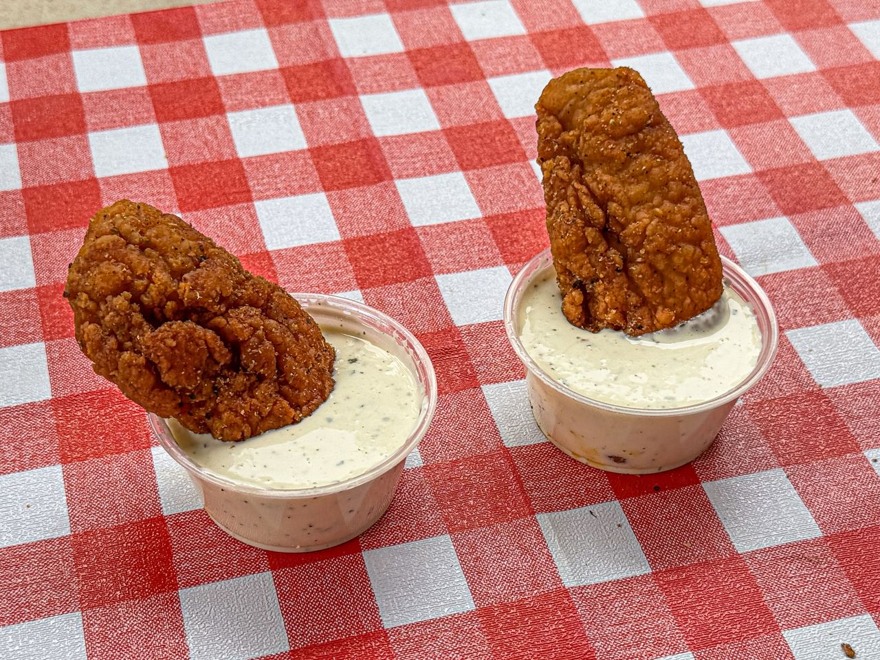 Two fried chicken tenders in individual cups of white dipping sauce on a red checkered tablecloth