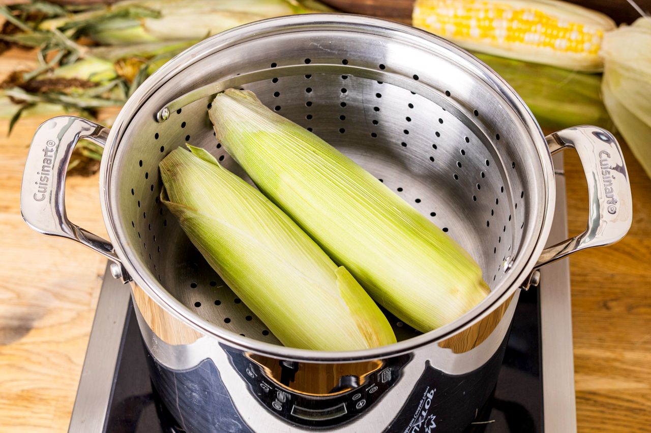 Corn in husks steaming in a pot on a stove kitchen setting with a wooden counter and corn in the background