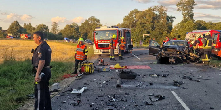 Verkehrsunfall mit Trümmern, Polizei und Feuerwehrleuten auf Landstraße bei Sonnenuntergang.