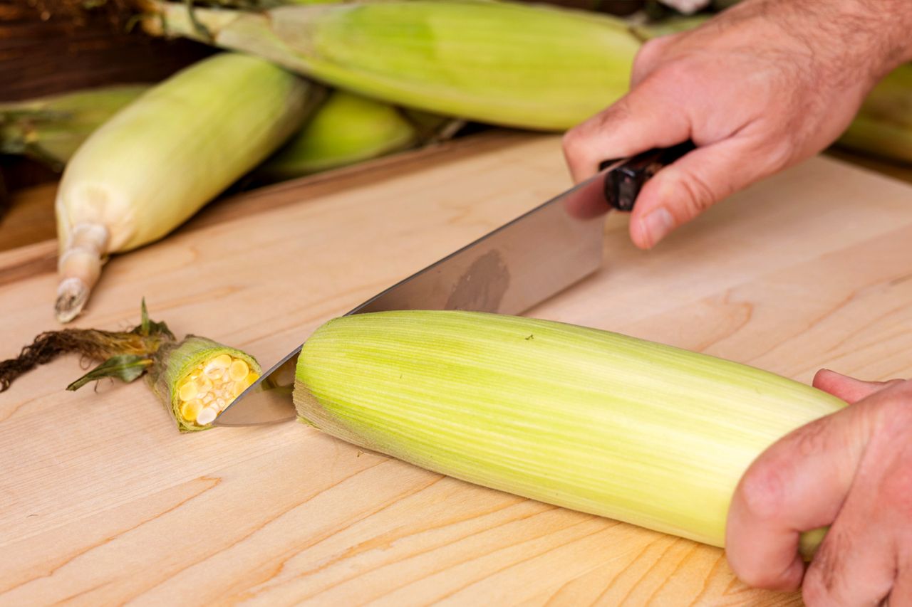 A person using a knife to cut the base of an ear of corn on a wooden surface
