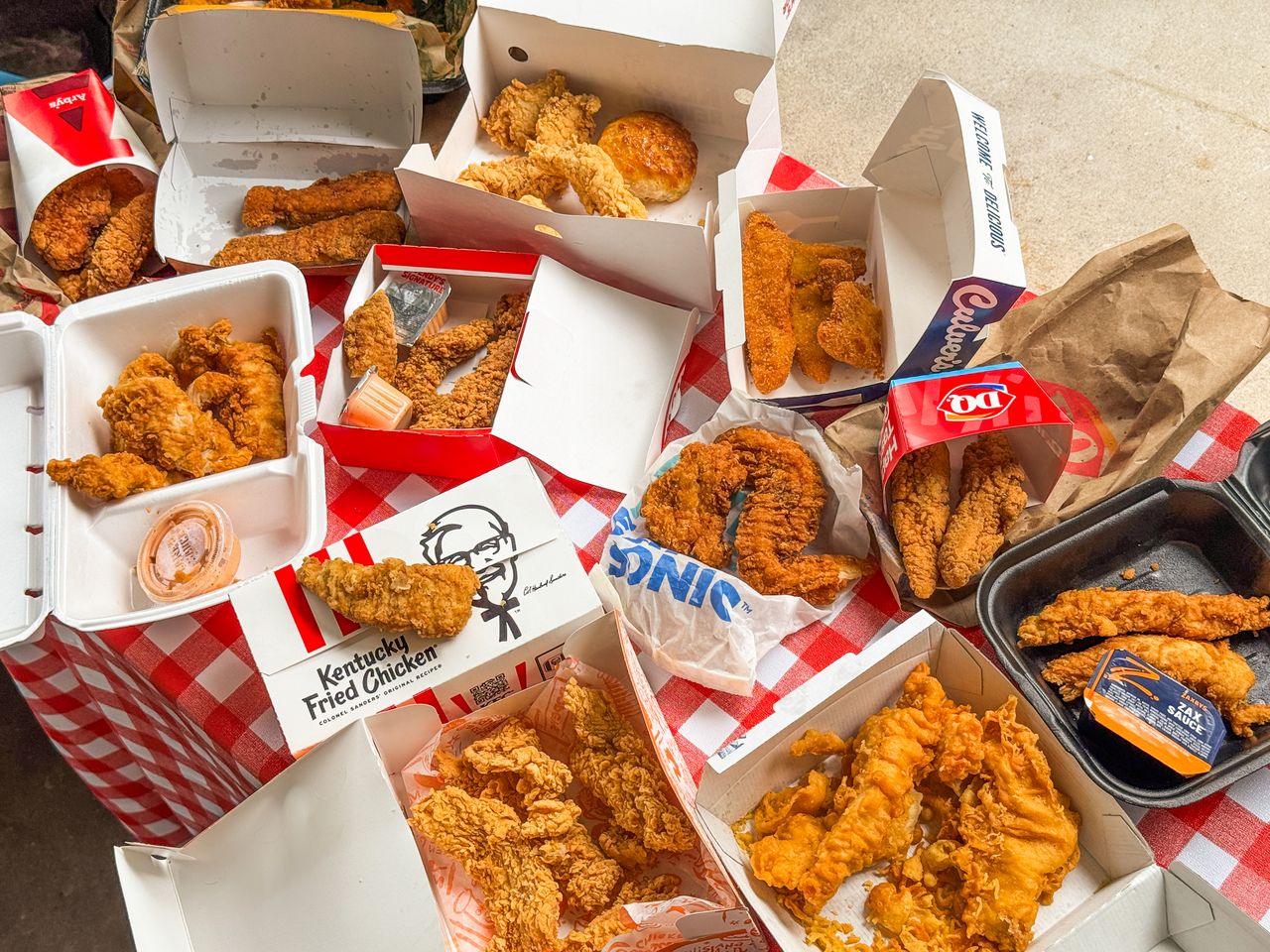 A variety of chicken tenders from multiple fastfood restaurants displayed on a checkered tablecloth