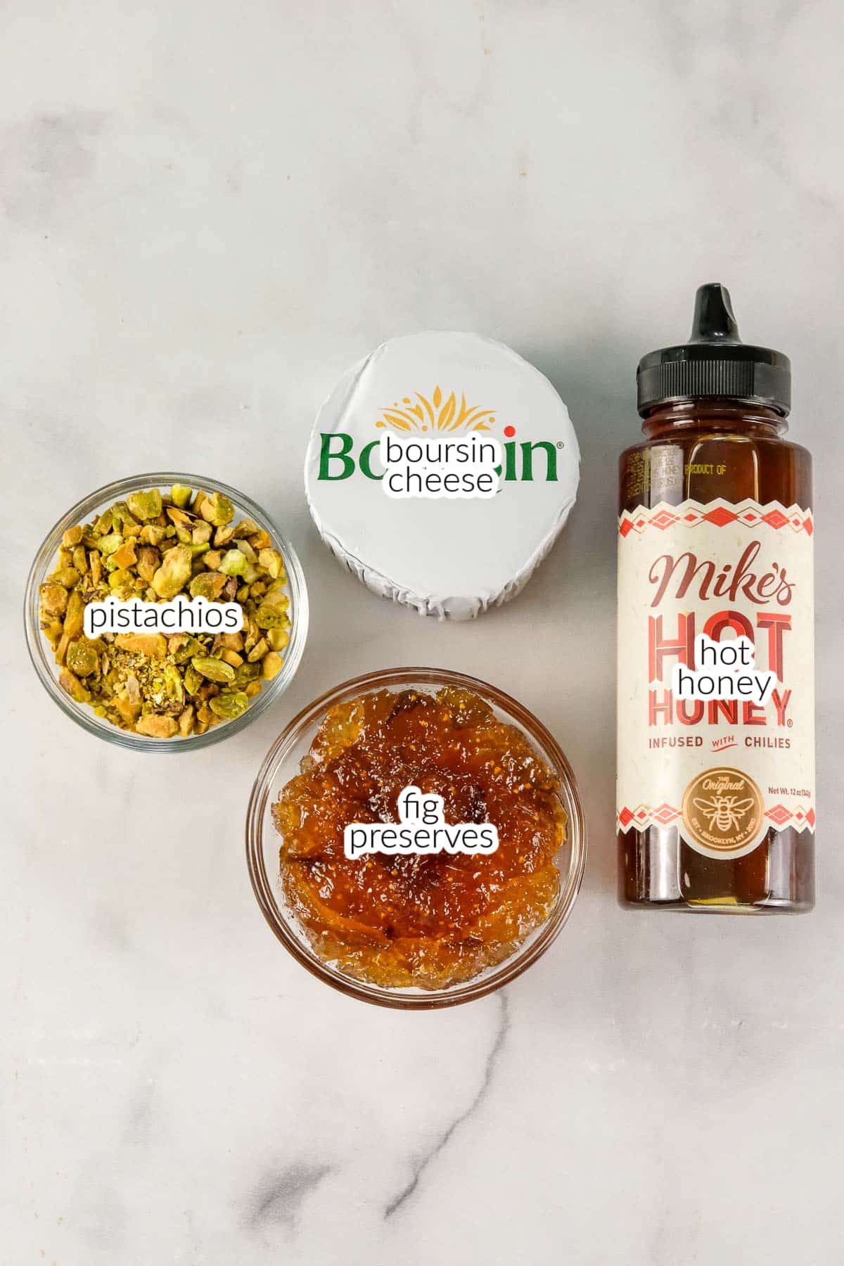 Ingredients for boursin cheese dip including a container of Boursin cheese, a bottle of Mike’s Hot Honey, a small bowl of chopped pistachios, and a small bowl of fig preserves sit on a white surface.