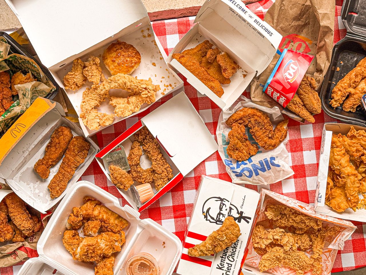 An assortment of chicken tenders and fastfood meals displayed on a checkered tablecloth featuring packaging from various brands