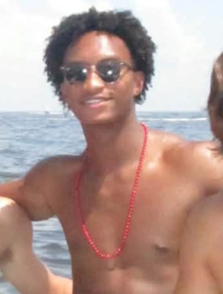 A person wearing sunglasses a red bead necklace and standing outdoors near a body of water