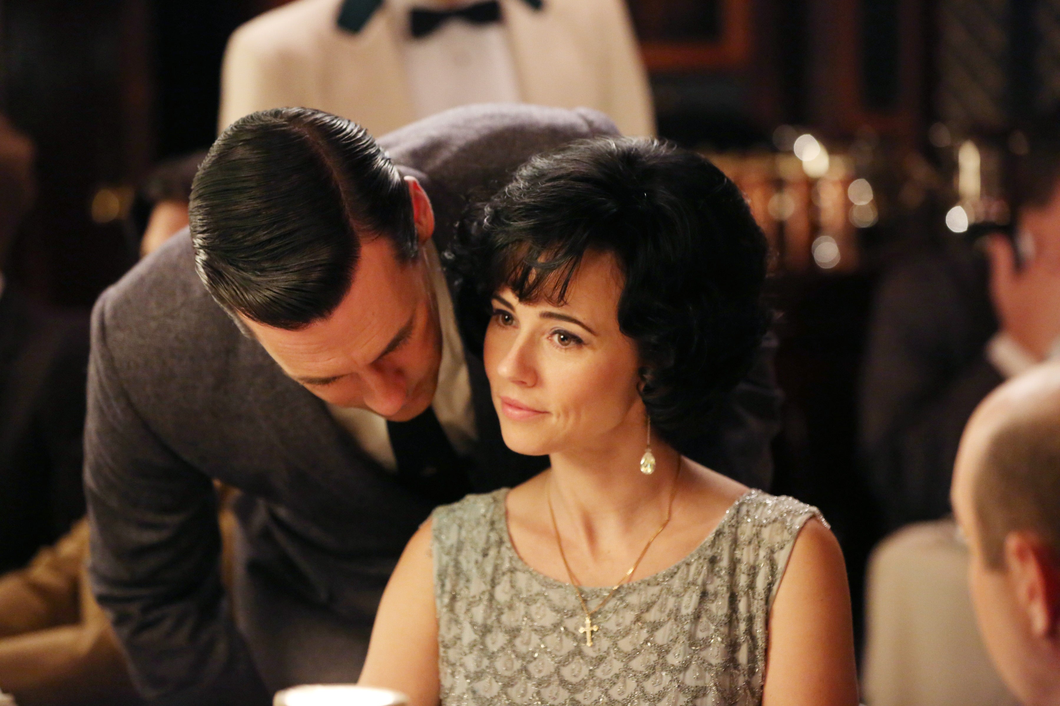 A person in formal attire leans in to speak with another person seated at a table in a restaurant setting