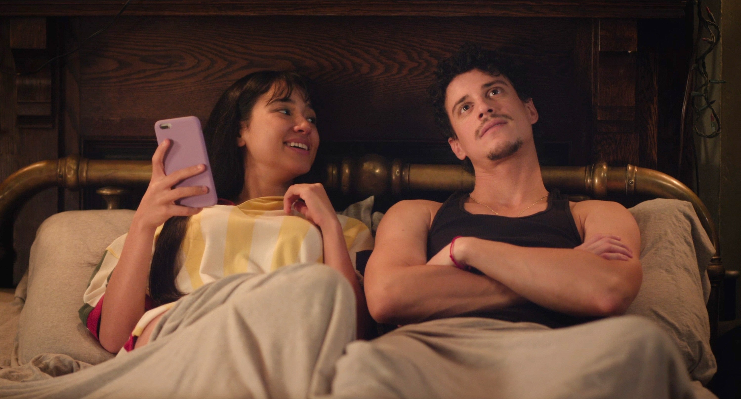 A couple relaxes in bed; one holds a phone, smiling, while the other looks contemplative. Cozy, intimate atmosphere