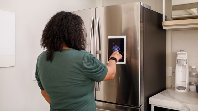 Person using a touchscreen panel on a stainless steel smart refrigerator in a kitchen.