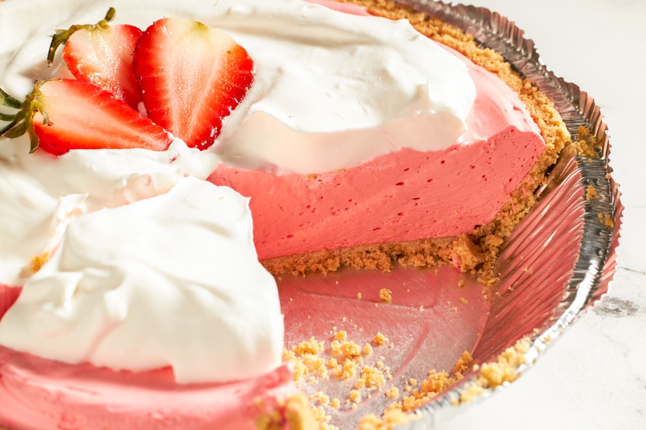 A sliced pie with a creamy pink filling whipped topping and strawberry garnish in a graham cracker crust