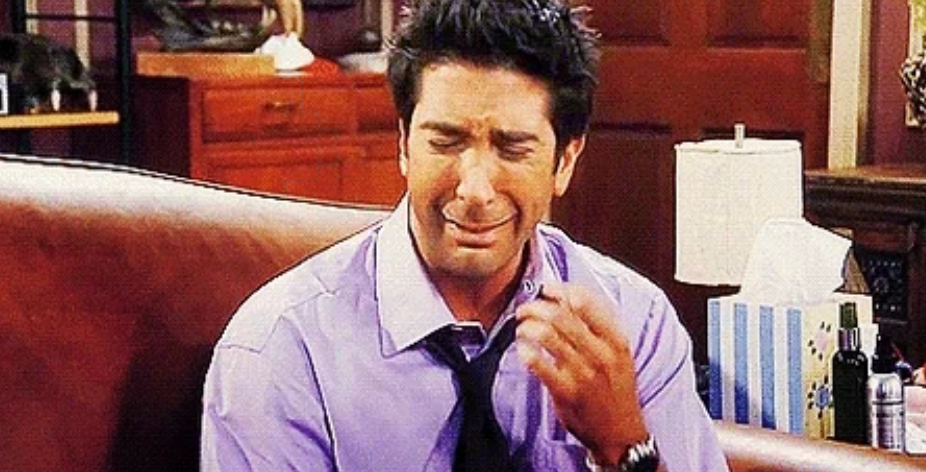 A person in a tie is sitting on a couch, visibly upset and crying, with a tissue box nearby, in a living room setting