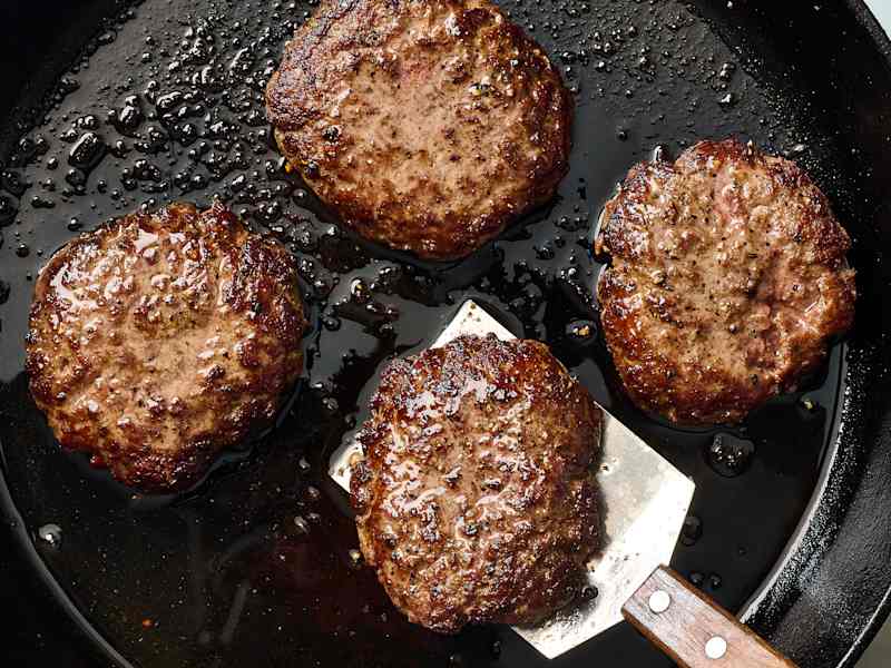 Four cooked hamburger patties in a skillet, glistening with oil, with a spatula lifting one.