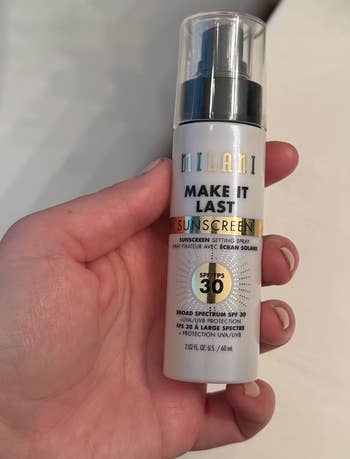 A hand holding a Milani Make It Last Sunscreen Setting Spray SPF 30 bottle, suitable for combining makeup setting and sun protection