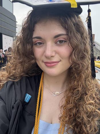 Person with curly hair in graduation attire smiles at the camera. Wearing a cap and gown with a gold cord and light blue top