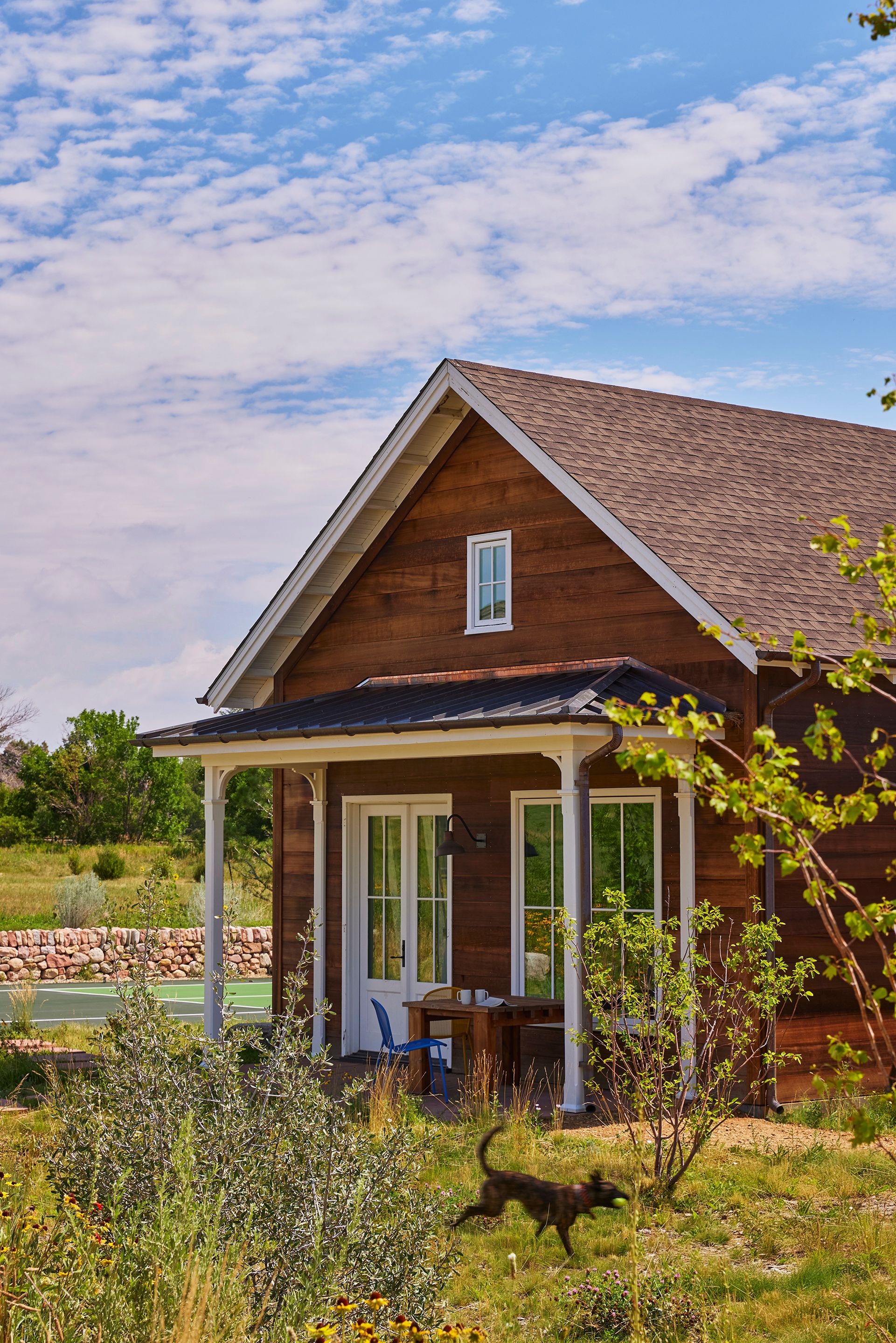 The guest cabin, directly adjacent to the primary house, is connected by a carport. The horizontal shiplap siding is clear red-wire brushed cedar sourced by Coburn Partners. The chairs are by Emu. The light is a Farmhouse gooseneck bracket in copper by Bevolo.