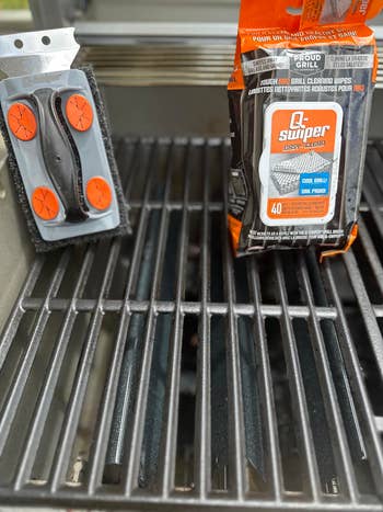 Grill cleaning kit on a grill, including a brush with orange pads and a pack of cleaning wipes for easy maintenance and care